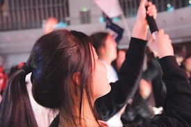 A young person with dark hair tied in a ponytail is seen in profile, holding an item that resembles a phone or a camera. The setting appears to be indoors, with other people blurred in the background, suggesting a crowded or lively environment.