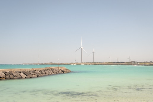 A serene coastal scene with turquoise waters and a rocky breakwater extending into the sea. In the background, multiple large wind turbines stand on a distant strip of land under a clear blue sky.