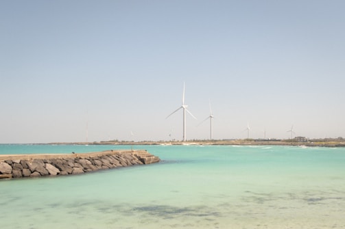A serene coastal scene with turquoise waters and a rocky breakwater extending into the sea. In the background, multiple large wind turbines stand on a distant strip of land under a clear blue sky.