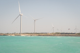 Several large wind turbines are standing on a strip of land with a clear blue sky above. The foreground features a vibrant turquoise ocean with slight waves. A few kites are visible in the sky, suggesting kite surfing activities near the shore.