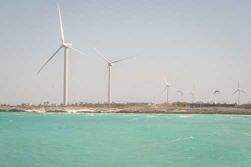 Several large wind turbines are standing on a strip of land with a clear blue sky above. The foreground features a vibrant turquoise ocean with slight waves. A few kites are visible in the sky, suggesting kite surfing activities near the shore.