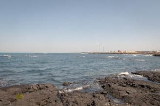 A rocky shoreline with waves crashing against the dark, jagged rocks. Wind turbines appear in the background, situated along the coastline. The sky is clear with a pale blue hue, and there are a few buildings visible in the distance.