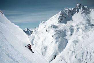 A snowboarder descends a steep, snow-covered mountain slope surrounded by rugged, majestic peaks. The skier is in motion, navigating the pristine white powder under a clear blue sky.