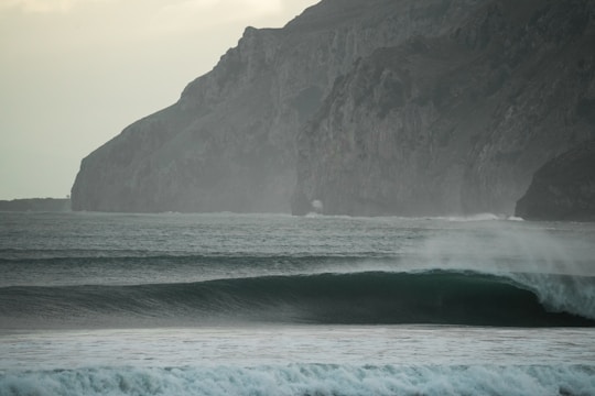 Dynamic ocean waves crashing against a rocky coastline under a cloudy sky.