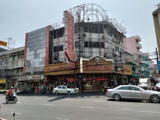 An urban street scene featuring a busy intersection with motorcycles and cars in the foreground. The background consists of multistory buildings, one of which is under construction, covered with scaffolding. The building displays a prominent red sign with intricate lettering. Various shops including a 7-Eleven are visible at street level, with signs in a language that appears to be Thai.