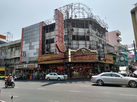 An urban street scene featuring a busy intersection with motorcycles and cars in the foreground. The background consists of multistory buildings, one of which is under construction, covered with scaffolding. The building displays a prominent red sign with intricate lettering. Various shops including a 7-Eleven are visible at street level, with signs in a language that appears to be Thai.