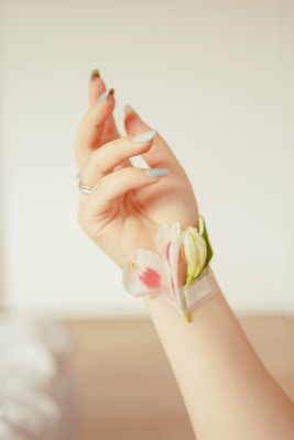A hand adorned with long, delicate fingernails holds a small arrangement of pink and white flowers nestled against the wrist. The nails are painted in a light, muted color, and the background is softly blurred, creating a gentle and serene atmosphere.