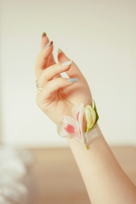 A hand adorned with long, delicate fingernails holds a small arrangement of pink and white flowers nestled against the wrist. The nails are painted in a light, muted color, and the background is softly blurred, creating a gentle and serene atmosphere.