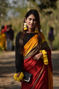 A woman is wearing a traditional sari with a combination of red and yellow. She has flowers in her hair and around her wrist. She is holding a black clutch purse and standing on a path in an outdoor setting with blurred greenery in the background.