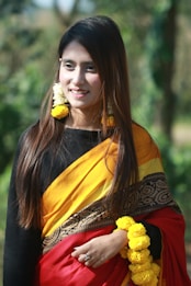 A woman with long hair is dressed in a vibrant traditional saree featuring shades of yellow, black, and red with intricate patterns. She accessorizes with yellow and white floral earrings and holds a garland of yellow flowers. The background is lush and blurred, suggesting an outdoor setting.