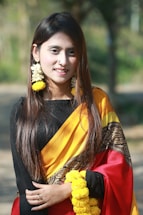 A woman with long, dark hair is wearing a traditional sari with a black top, yellow and red draped fabric, and intricate patterns. She is adorned with yellow floral jewelry, including earrings and bracelets, and has a small nose ring. The background is a blurred outdoor setting with greenery.