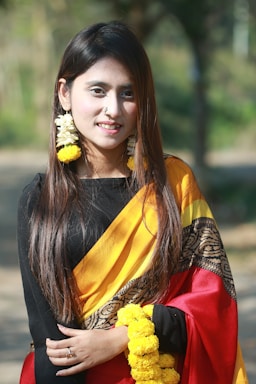 A woman with long, dark hair is wearing a traditional sari with a black top, yellow and red draped fabric, and intricate patterns. She is adorned with yellow floral jewelry, including earrings and bracelets, and has a small nose ring. The background is a blurred outdoor setting with greenery.