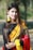 A woman with long, dark hair is wearing a traditional sari with a black top, yellow and red draped fabric, and intricate patterns. She is adorned with yellow floral jewelry, including earrings and bracelets, and has a small nose ring. The background is a blurred outdoor setting with greenery.