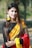 A woman with long, dark hair is wearing a traditional sari with a black top, yellow and red draped fabric, and intricate patterns. She is adorned with yellow floral jewelry, including earrings and bracelets, and has a small nose ring. The background is a blurred outdoor setting with greenery.