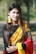 A woman with long, dark hair is wearing a traditional sari with a black top, yellow and red draped fabric, and intricate patterns. She is adorned with yellow floral jewelry, including earrings and bracelets, and has a small nose ring. The background is a blurred outdoor setting with greenery.