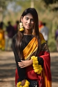 A person wearing a traditional colorful sari stands outdoors, adorned with bright yellow floral garlands. The sari features bold yellow and red hues with intricate patterns. The background is blurred with several people walking, suggesting an outdoor setting.