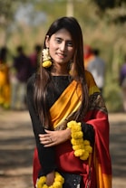 A person wearing a traditional colorful sari stands outdoors, adorned with bright yellow floral garlands. The sari features bold yellow and red hues with intricate patterns. The background is blurred with several people walking, suggesting an outdoor setting.