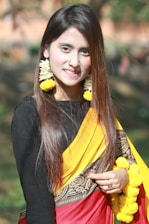 A woman with long brown hair is wearing a traditional saree with a red, yellow, and black design. She accessorizes with large garlands of yellow flowers as earrings and holds more flowers in her hand. She has a nose ring and poses outdoors in a natural setting with a blurred green background.