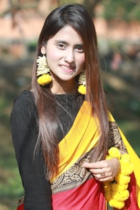 A woman with long brown hair is wearing a traditional saree with a red, yellow, and black design. She accessorizes with large garlands of yellow flowers as earrings and holds more flowers in her hand. She has a nose ring and poses outdoors in a natural setting with a blurred green background.