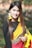 A woman with long brown hair is wearing a traditional saree with a red, yellow, and black design. She accessorizes with large garlands of yellow flowers as earrings and holds more flowers in her hand. She has a nose ring and poses outdoors in a natural setting with a blurred green background.