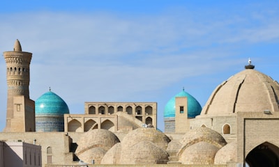 A skyline dominantly featuring a series of mosque buildings with intricate architecture. The structures display large domes in vibrant turquoise and earthy tones beneath a bright blue sky. The ornate minaret adds a historical and cultural aspect to the setting.