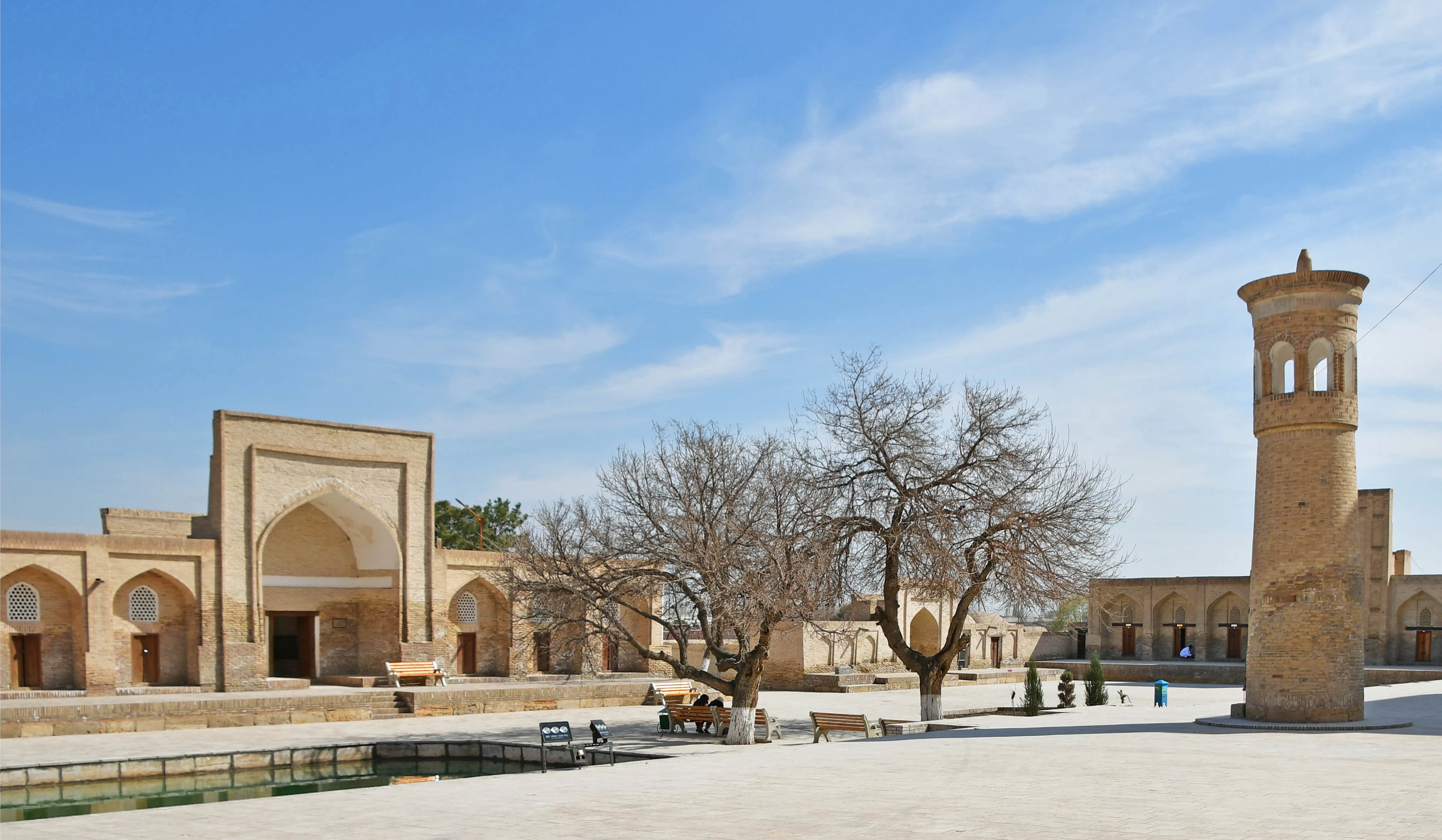 The wide sunlit courtyard of the Chor-Bakr necropolis outside Bukhara, its low arcaded brick galleries and slender minaret standing around a still green pool with bare trees and benches.