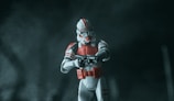 A toy figurine of a soldier is displayed, wearing a white and red suit of armor and holding a blaster. The background is dark and misty, creating a mysterious and dramatic atmosphere.