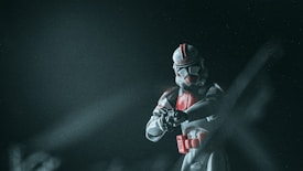 A toy figure resembling a sci-fi soldier is centered against a dark, misty background. The figure is clad in white armor with red accents, holding a weapon in a poised stance. There is a sense of movement from the blurred elements in the foreground.