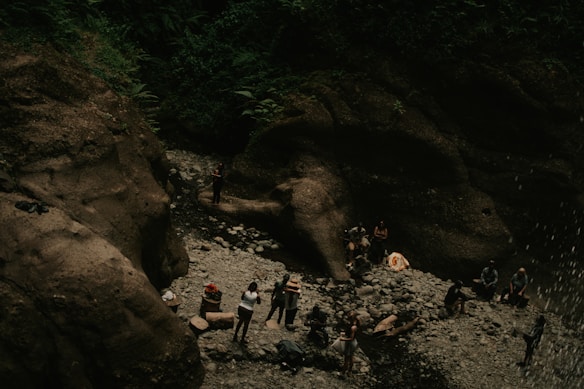 A group of people is gathered at the base of a rock formation in a forested area. They are situated near a rocky terrain with some boulders and appear to be engaged in various activities. The area is surrounded by dense green foliage, creating a natural and serene environment.