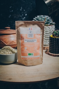 A bag of shelled hemp seeds is prominently displayed on a wooden table. The packaging is made of brown paper and features labels in French, highlighting its organic certification and vegan nature. Surrounding the bag are ceramic containers and small potted cacti, adding a natural and rustic feel to the scene.