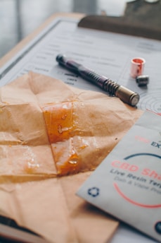 A close-up view of a piece of parchment paper with a sticky substance, possibly resin or concentrate, on top. Next to it is a CBD Shatter packaging labeled 'Live Resin Terpenes'. A clipboard with some documents and a pen with a metallic tip are visible in the background.