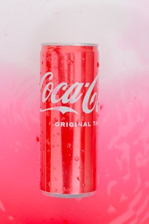 A red can with the brand name printed in white letters, surrounded by water droplets on its surface, sits against a pink and white gradient background.
