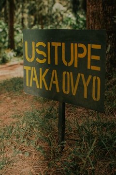 A green metal sign with yellow lettering is placed in a grassy area, partially shaded by a tree. The sign bears a message written in a bold, straightforward font.