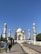 A grand white mausoleum with intricate architectural details, featuring four minarets and a large dome, surrounded by well-maintained gardens. A group of visitors is present, walking along a wide pathway leading to the structure.