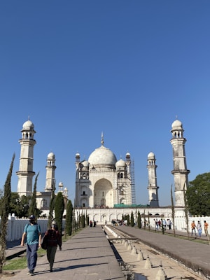 A grand white mausoleum with intricate architectural details, featuring four minarets and a large dome, surrounded by well-maintained gardens. A group of visitors is present, walking along a wide pathway leading to the structure.