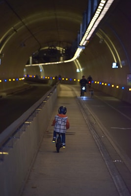 A child wearing a helmet rides a small bicycle along a designated bike lane inside a dimly lit tunnel. The tunnel is illuminated by a series of small, evenly spaced yellow and blue lights along the walls, creating a modern and secure atmosphere. In the distance, two more cyclists can be seen approaching, also using the bike lane.