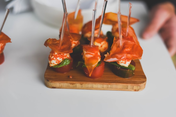 A selection of appetizers arranged on a wooden serving board. The skewered bites consist of mozzarella balls, cherry tomato halves, basil leaves, and slices of cured meat. The colorful presentation is enhanced by the bright red and green ingredients, creating an inviting appearance.
