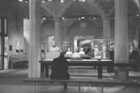A person sits on a bench in a museum, facing a display of classical sculptures and artifacts. The room is adorned with stone columns and arches, creating an ancient and historical ambiance. Busts and statues are encased in glass or displayed on pedestals, surrounded by soft lighting that highlights their details.