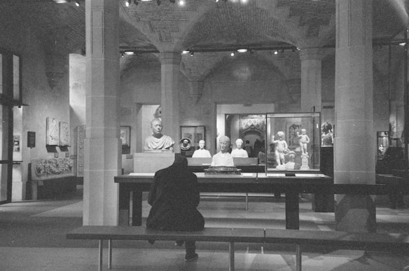 A person sits on a bench in a museum, facing a display of classical sculptures and artifacts. The room is adorned with stone columns and arches, creating an ancient and historical ambiance. Busts and statues are encased in glass or displayed on pedestals, surrounded by soft lighting that highlights their details.