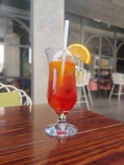 A tall glass filled with a colorful beverage featuring red and orange hues, garnished with a slice of lemon and a straw, is placed on a polished wooden table. The background consists of out-of-focus chairs and a large window, suggesting an indoor or patio setting.