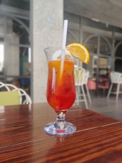 A tall glass filled with a colorful beverage featuring red and orange hues, garnished with a slice of lemon and a straw, is placed on a polished wooden table. The background consists of out-of-focus chairs and a large window, suggesting an indoor or patio setting.