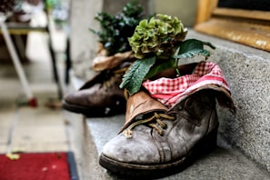 A pair of worn-out, brown leather boots is placed on stone steps, creatively repurposed as planters. One boot contains vibrant green foliage with a cluster of hydrangea flowers, while the interior is lined with red and white checkered fabric, adding a rustic charm.