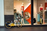 A storefront features mannequins dressed in colorful clothing, with a striking orange and teal display. Various accessories are arranged on shelves behind the window. A bicycle with a yellow accent is parked in front of the window.
