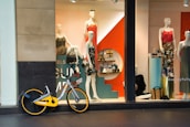 A storefront features mannequins dressed in colorful clothing, with a striking orange and teal display. Various accessories are arranged on shelves behind the window. A bicycle with a yellow accent is parked in front of the window.