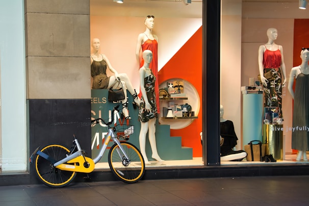 A storefront features mannequins dressed in colorful clothing, with a striking orange and teal display. Various accessories are arranged on shelves behind the window. A bicycle with a yellow accent is parked in front of the window.
