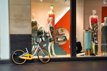 A storefront features mannequins dressed in colorful clothing, with a striking orange and teal display. Various accessories are arranged on shelves behind the window. A bicycle with a yellow accent is parked in front of the window.