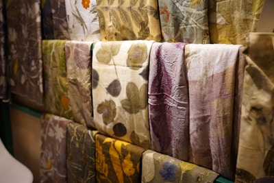 Several pieces of fabric or textile are displayed with natural patterns, featuring floral and leaf motifs in various muted colors. The arrangement showcases an artisanal style with each piece exhibiting unique designs and textures.