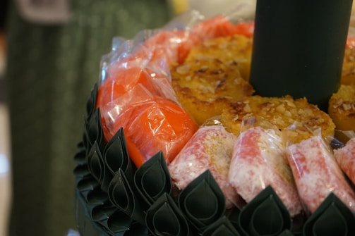 Assorted sweets are arranged in a circular pattern, wrapped in plastic. The sweets include yellow and red colored pieces, possibly indicating different flavors or types. They sit on a decorative green base with a leaf-like texture, suggesting a festive or special occasion presentation.