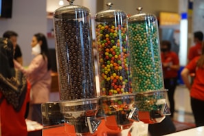 Three tall, transparent dispensers are filled with colorful candy or snack pellets. The left dispenser contains chocolate or dark-colored spherical snacks, the middle is filled with vibrant and multicolored candy, and the right one holds turquoise and light green-colored spheres. In the background, several people are present, some wearing masks, against a blurred indoor setting.