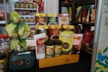 A display of various packaged food products is arranged on a wooden platform. The products include snack packs and containers in vibrant packaging with different colors such as green, yellow, and red. A decorative item resembling a small tree with gold and green leaves is placed alongside. Additionally, there is a small, dark green retro-style radio on the left side of the arrangement.